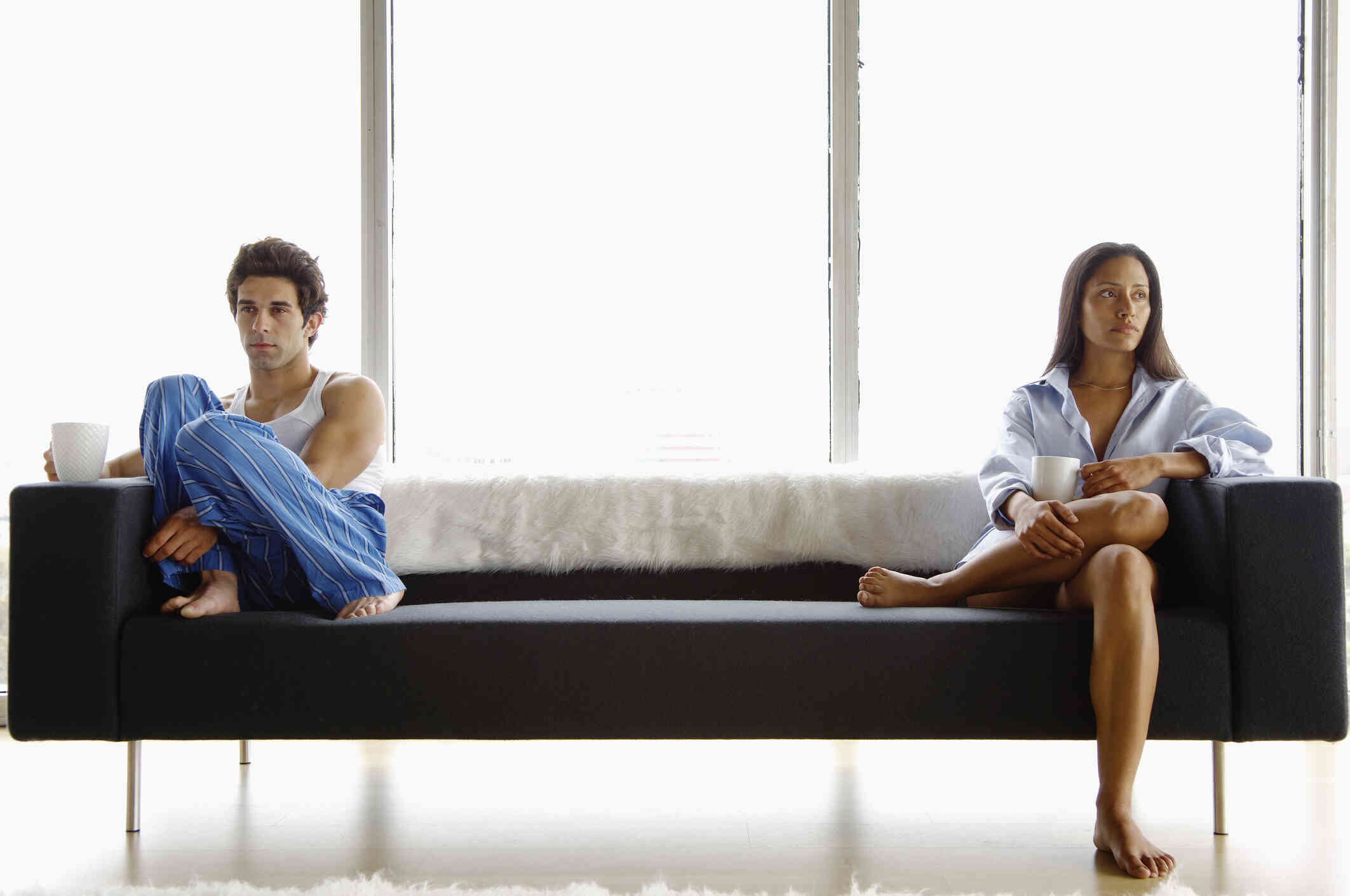 A male and female couple in pajamas sit on opposite ends of a couch while looking upset.